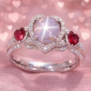 Può includere: Un anello d'argento con una gemma a forma di cuore, di colore viola chiaro, al centro, circondata da un alone di piccoli diamanti. Due gemme rosse a forma di cuore affiancano la pietra centrale. La fascia è anch'essa incastonata con piccoli diamanti.