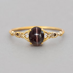 Op de afbeelding: Een gouden ring met een ovale, donkerpaarse ster-edelsteen in een klauwzetting. De ringband is voorzien van gedetailleerd metaalwerk met decoratieve krulpatronen aan beide zijden van de centrale steen, wat zorgt voor een verfijnde uitstraling.