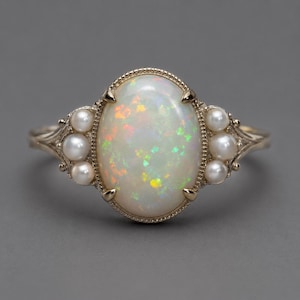 Può includere: Anello in oro con un'opale ovale che mostra lampi iridescenti. L'anello è impreziosito da piccole perle rotonde su entrambi i lati dell'opale. L'incastonatura presenta un design delicato di ispirazione vintage.