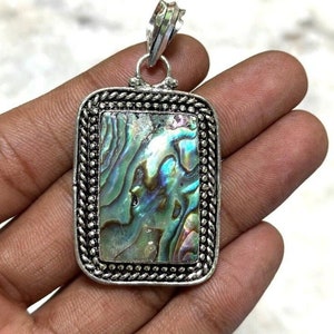 Può includere: Un ciondolo d'argento con un'incisione rettangolare di madreperla di abalone. La conchiglia presenta colori iridescenti di blu, verde e viola. Il ciondolo ha un bordo d'argento dettagliato.