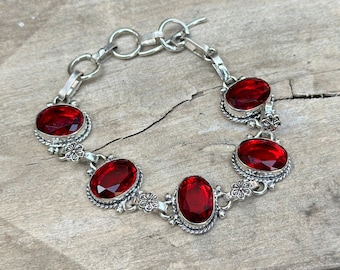 May include: A silver bracelet featuring oval-cut, deep red gemstones. The gemstones are set in ornate, floral-inspired bezels. The bracelet has a toggle clasp and is displayed on a wooden surface. The bracelet is a piece of jewelry.
