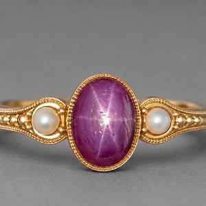 Pode incluir: Um anel de ouro com um rubi estrela central em corte cabochão oval, exibindo um efeito de estrela de seis pontas. O rubi é ladeado por duas pequenas pérolas brancas redondas, montadas numa banda de ouro com detalhes metálicos em relevo.