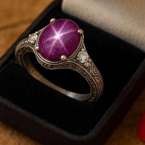 Puede incluir: Anillo de plata con una gema de rubí estrella ovalada. El rubí es de color magenta intenso con un efecto de estrella de seis rayos. El anillo tiene intrincados detalles de filigrana y dos pequeñas gemas claras a cada lado del rubí.