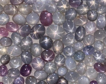 May include: A collection of star sapphire cabochons in various shades of gray and purple. The stones are all oval shaped and have a star-like pattern in the center.