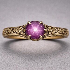 Op de afbeelding: Een gouden ring met een grote, ronde, paarsrode edelsteen met een ster-effect. De band is versierd met bloemmotieven. De ring staat tegen een grijze achtergrond.
