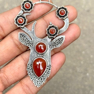 Puede incluir: Pendiente de plata con forma de silueta de ciervo con detalles intrincados. El ciervo tiene cinco piedras de cornalina roja engastadas en sus astas y dos piedras de cornalina más grandes engastadas en su cuerpo.
