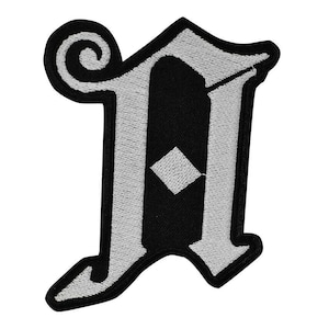 Könnte beinhalten: Ein schwarz-weißer, gestickter Aufnäher in Form des Buchstabens "N". Der Buchstabe hat einen gotischen Schriftstil mit weißem Umriss und schwarzer Mitte. Der Aufnäher hat eine dekorative Spirale oben.