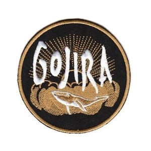 May include: Embroidered patch featuring the word "GOJIRA" in white script above a whale illustration. The design is set on a black circular background with a gold border and accents. A unique patch for jackets, bags, or other apparel.