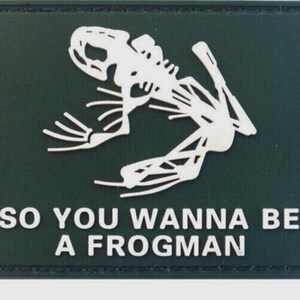 May include: A rectangular dark green patch featuring a white graphic of a frog skeleton in a diving pose. Below the skeleton, the text reads SO YOU WANNA BE A FROGMAN in white capital letters. The patch has a stitched border and a matte finish.