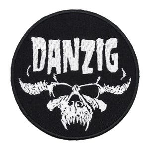 Może przedstawiać: Okrągła, czarna naszywka z białym napisem "DANZIG" w stylizowanej czcionce. Poniżej tekstu znajduje się biały wzór czaszki z rogami. Naszywka jest prawdopodobnie haftowana lub drukowana, odpowiednia do umieszczenia na odzieży lub akcesoriach.