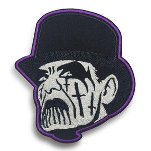 May include: An embroidered patch featuring a black top hat with a purple border. The design depicts a white face with dark, heavy makeup, including vertical cross symbols under the eyes and dark, dripping lips, set against a black background.