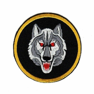 May include: A round embroidered patch with a black background and a gold border. The patch features a white wolf with red eyes and a snarling expression.