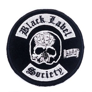 May include: A round, black patch with white embroidered text and a skull design. The text reads "Black Label Society" in an arch, with "Society" at the bottom. A banner with text is on the right side.