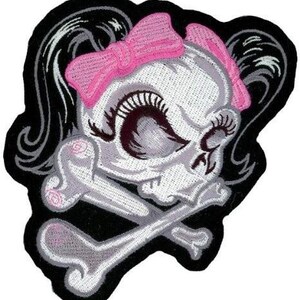 May include: An embroidered patch featuring a stylised skull with long black hair, a bright pink bow, and prominent eyelashes. The skull is positioned over two crossed bones, all outlined in black thread against a solid black background.