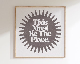 May include: A framed print with a brown sunburst design and the text "This Must Be The Place." in white.