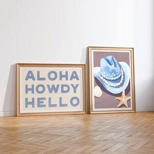 May include: Two framed prints with a light beige background. The print on the left says "ALOHA HOWDY HELLO" in blue letters. The print on the right shows a blue cowboy hat with white stars and a pearl necklace on a brown background. A starfish and a seashell are also on the background.