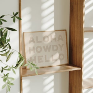 Może przedstawiać: Wydruk w ramie z napisem "Aloha Howdy Hello" w jasnobrązowym kolorze na białym tle. Wydruk znajduje się na drewnianej półce z zieloną rośliną w tle.