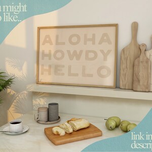 Puede incluir: Una impresi&oacute;n enmarcada con el texto "ALOHA HOWDY HELLO" en color marr&oacute;n claro sobre un fondo beige. La impresi&oacute;n est&aacute; en un estante blanco con dos tablas de cortar de madera.