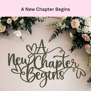 May include: A decorative sign with the phrase "A New Chapter Begins" in a cursive font. The sign is dark green and set against a light background, with floral arrangements of roses and greenery framing the top.