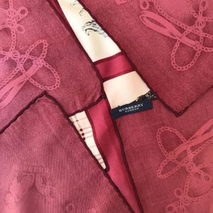 May include: A collection of pink handkerchiefs with a decorative design and dark red borders. A section of a patterned scarf with the text "BURBERRY LONDON" is visible, adding a touch of luxury to the arrangement. The scarf features a cream background with horse and rider illustrations.
