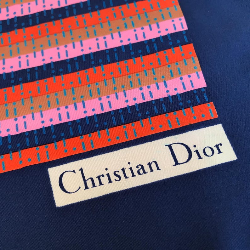 May include: A navy blue silk scarf with a colorful striped pattern. The stripes are in shades of orange, pink, brown, and blue, with exclamation point accents. The words "Christian Dior" are printed in a white rectangle.