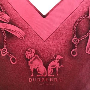 May include: A close-up of a pink scarf with a V-neck design. The scarf features a repeating pattern of dog silhouettes, chains, and the text "BURBERRY LONDON". The scarf has a textured appearance.