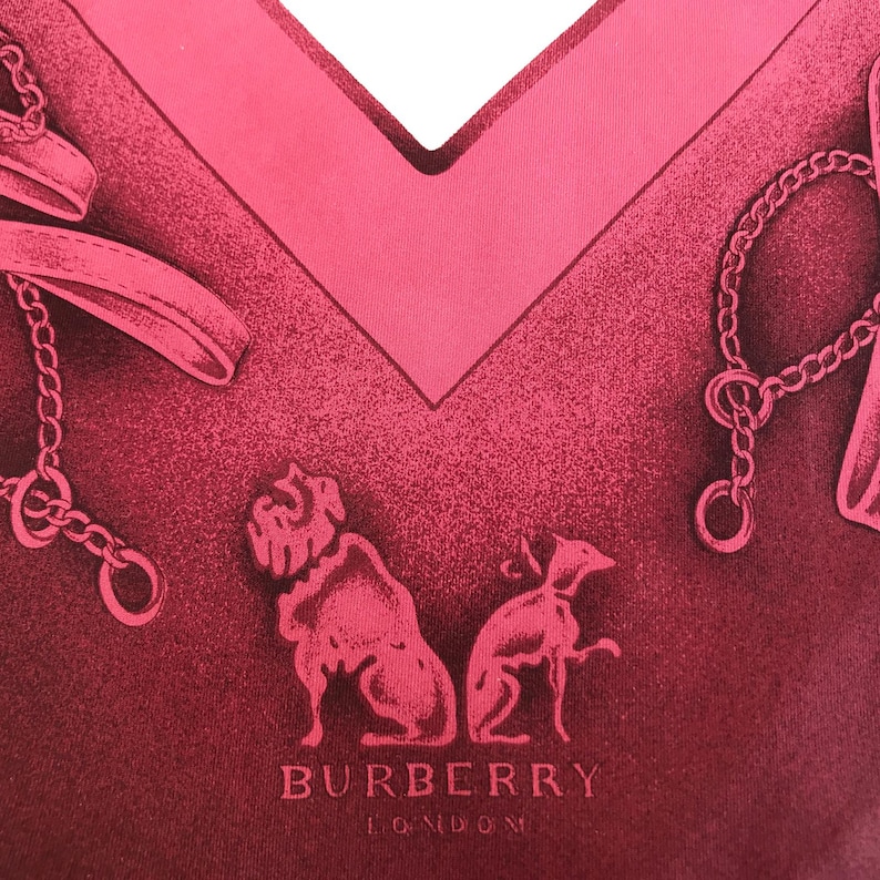 May include: A close-up of a pink scarf with a V-neck design. The scarf features a repeating pattern of dog silhouettes, chains, and the text "BURBERRY LONDON". The scarf has a textured appearance.