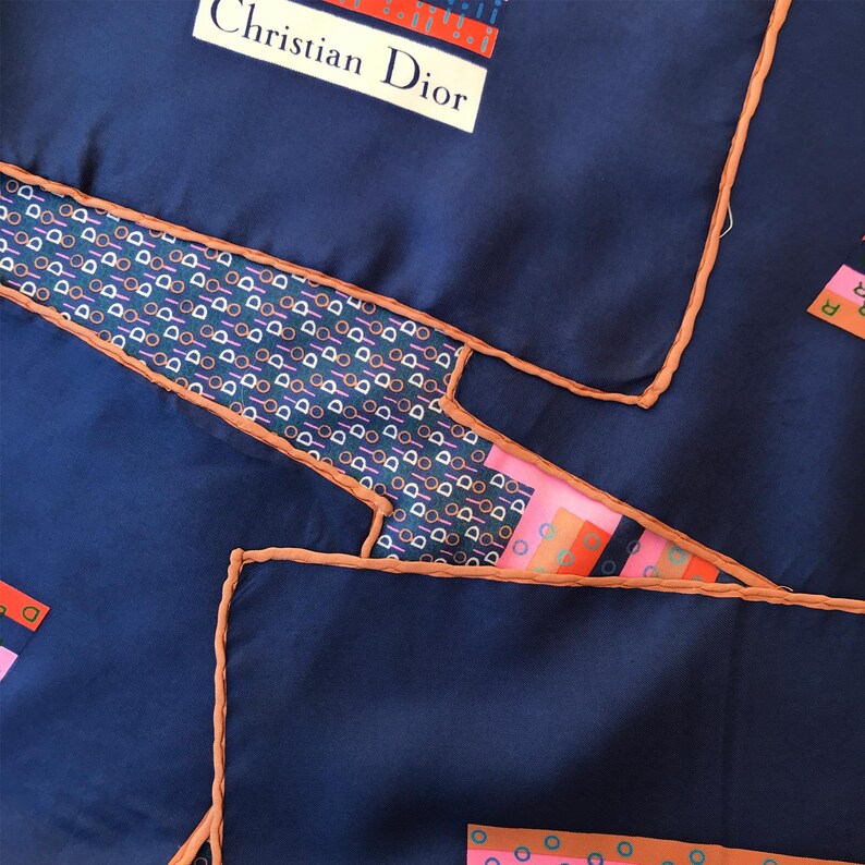 May include: A navy blue silk scarf with the text "Christian Dior" in white. The scarf features geometric patterns in pink, orange, and blue, with a contrasting orange border. The design includes repeating patterns of circles and lines.
