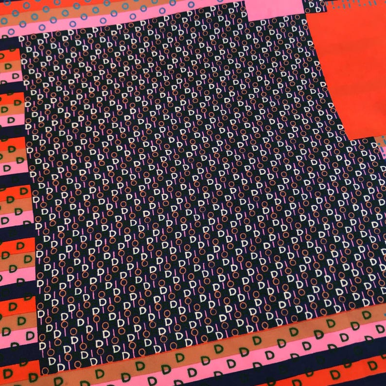 May include: A patterned textile featuring a repeating design of the letter "D" and circles in white, pink, and orange against a dark background. The design includes stripes and solid color blocks in pink, orange, and navy blue.