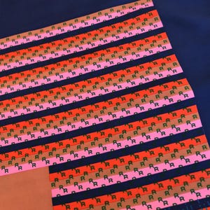 May include: A patterned textile featuring rows of the letter "R" in black, arranged on stripes of red, pink, and brown. The background is a dark blue. The design is repeated across the fabric.