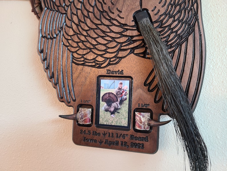 K&ouml;nnte beinhalten: Ein h&ouml;lzerner Truthahn-Wandhalter mit einem Foto eines J&auml;gers und eines Truthahns. Der Halter ist mit dem Text "David", "24.3 lbs", "11 1/4" Beard", "Iowa" und "April 12, 2021" graviert.