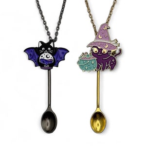 May include: Two black cat themed spoon necklaces. One features a black cat with bat wings holding a potion, the other features a black cat wearing a purple witch hat holding a cauldron. Both spoons are silver and gold.