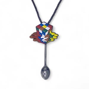 Op de afbeelding: Een zwarte bolo tie met een zilveren lepelscharm. De lepel heeft een kleurrijk emaille ontwerp van een smeltende Rubik's Cube.