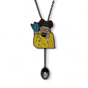 Puede incluir: Un collar de metal negro con una imagen de dibujos animados de un personaje que lleva un mono amarillo y una máscara de gas, sosteniendo un guante azul. El personaje sostiene una cuchara con un cristal azul en su interior.