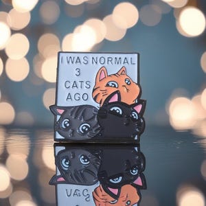 May include: A rectangular enamel pin with the text "I WAS NORMAL 3 CATS AGO." The pin features three cat faces: orange tabby, black, and gray. The pin is reflected on a shiny surface.