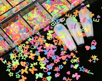 May include: A clear plastic container with 12 compartments filled with colourful glitter and confetti. The glitter and confetti are in the shapes of hearts, stars, butterflies, and circles. The container is open and the glitter and confetti are spilling out onto a black surface.