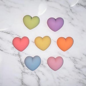May include: Seven colorful heart-shaped objects in shades of green, purple, red, yellow, orange, blue, and pink are arranged on a white marble surface. The hearts are smooth and have a matte finish. The image is well-lit, highlighting the colors.