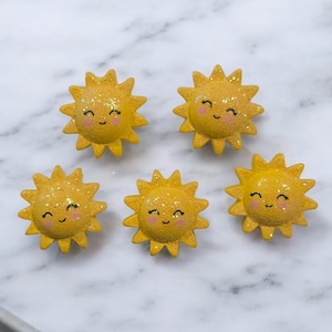 May include: Five yellow sun-shaped decorative embellishments with glitter and smiling faces. The suns have a textured surface and are arranged on a white marble background. The suns are a cheerful, whimsical design.