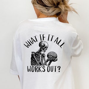 Op de afbeelding: Wit T-shirt met een zwart-witte afbeelding van een skelet dat een hersenen vasthoudt. De tekst "What if it all works out?" staat op het shirt gedrukt.