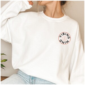 Puede incluir: Sudadera blanca con un c&iacute;rculo rosa y texto negro que dice "VIBING MIND". Dentro del c&iacute;rculo hay un gr&aacute;fico de cerebro rosa.
