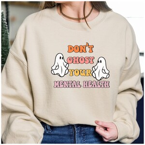 May include: A cream colored sweatshirt with the text "Don't Ghost Your Mental Health" in pink, orange, and white letters. Two white ghosts are on either side of the text.