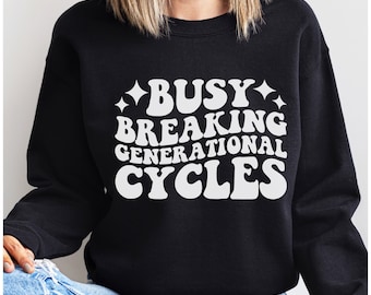 Op de afbeelding: Zwart sweatshirt met witte tekst die "Busy Breaking Generational Cycles" in een retro-lettertype zegt.