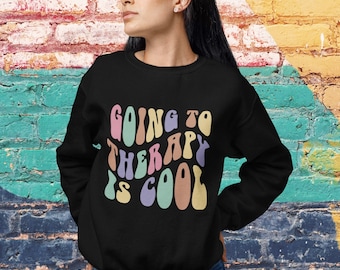 Op de afbeelding: Een zwart sweatshirt met de tekst "Going to Therapy is Cool" in een retro, kleurrijk lettertype.