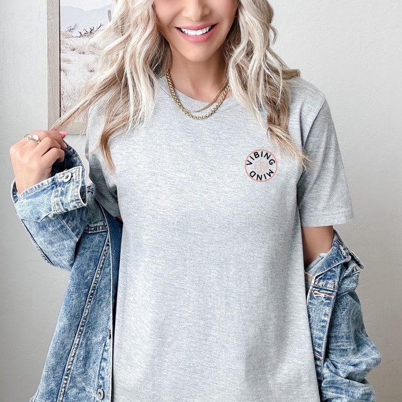 Op de afbeelding: Een grijs T-shirt met een zwarte cirkelvormige afbeelding met de tekst "Vibing Mind" in wit.