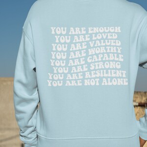 May include: A light blue sweatshirt with the text "YOU ARE ENOUGH YOU ARE LOVED YOU ARE VALUED YOU ARE WORTHY YOU ARE CAPABLE YOU ARE STRONG YOU ARE RESILIENT YOU ARE NOT ALONE" printed in white on the back.