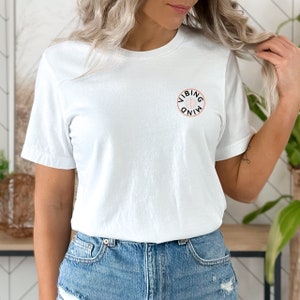 Op de afbeelding: Wit T-shirt met een zwarte cirkelvormige afbeelding met de tekst "VIBING MIND" in wit.