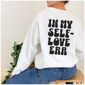 Puede incluir: Sudadera blanca con texto negro que dice "In My Self-Love Era" en una fuente retro.