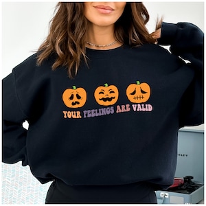 Peut inclure: Sweat-shirt noir avec un graphique de trois citrouilles orange aux expressions faciales différentes. Le texte "YOUR FEELINGS ARE VALID" est imprimé sous les citrouilles.