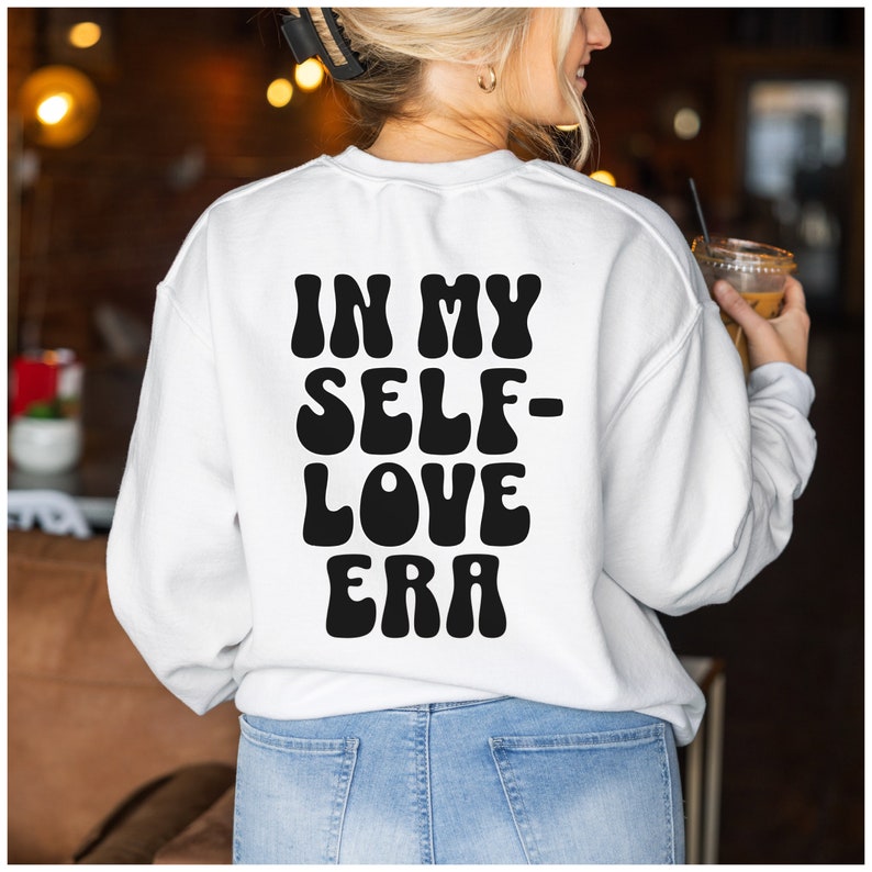 Puede incluir: Sudadera blanca con texto negro que dice "In My Self-Love Era" en una fuente retro.