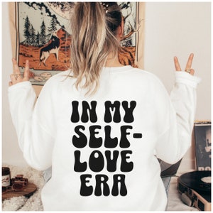 Puede incluir: Sudadera blanca con texto negro que dice "In My Self-Love Era" en una fuente retro.